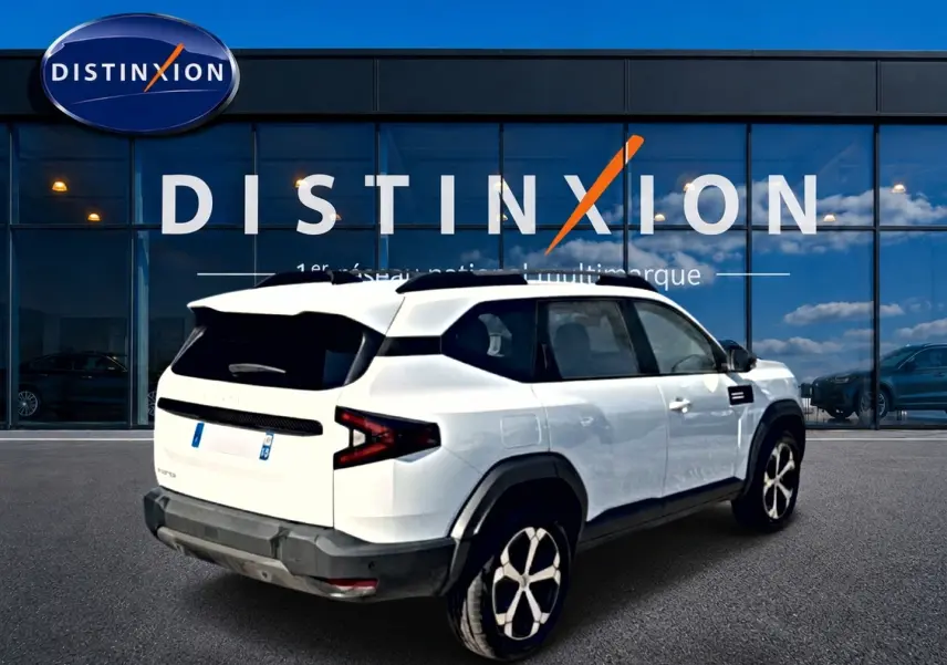 Vue 3/4 arrière droite du SUV blanc Dacia Bigster 2026 avec jantes alu et toit noir devant une vitrine Distinxion.