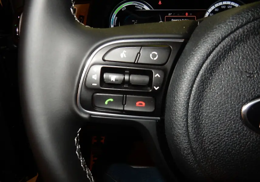 Gros plan sur les commandes gauche du volant noir du Kia Niro hybride gris galène, avec tableau de bord numérique en arrière-plan.