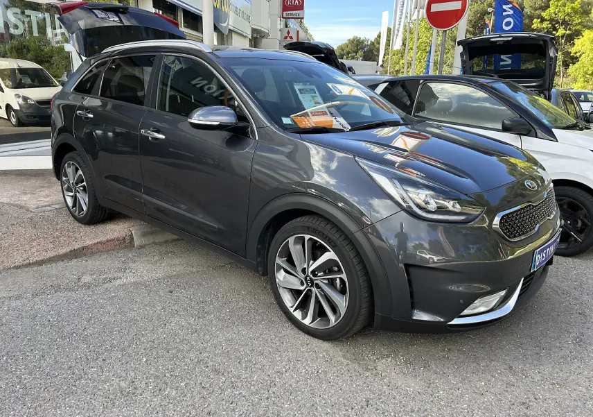 KIA Niro hybride gris galène en 3/4 avant droit, avec jantes alliage et feux LED visibles, coffre arrière ouvert.