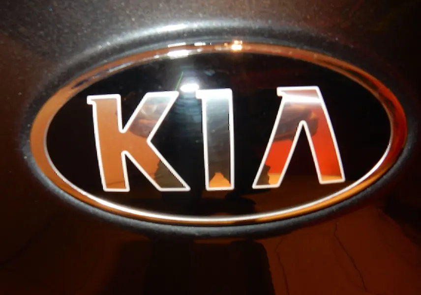 Gros plan sur le logo KIA en chrome sur fond gris galène métallique, éclairé par une lumière chaude.