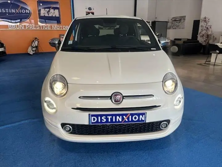 Vue avant d'une FIAT 500 blanche avec phares allumés, exposée en showroom sur sol bleu.