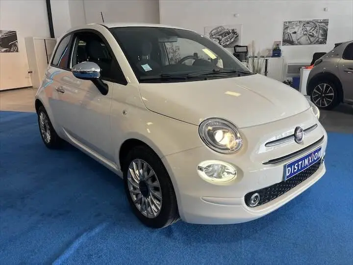 Vue 3/4 avant d'une FIAT 500 blanche 2023 avec phares allumés, jantes alliage et rétroviseurs chromés en showroom.