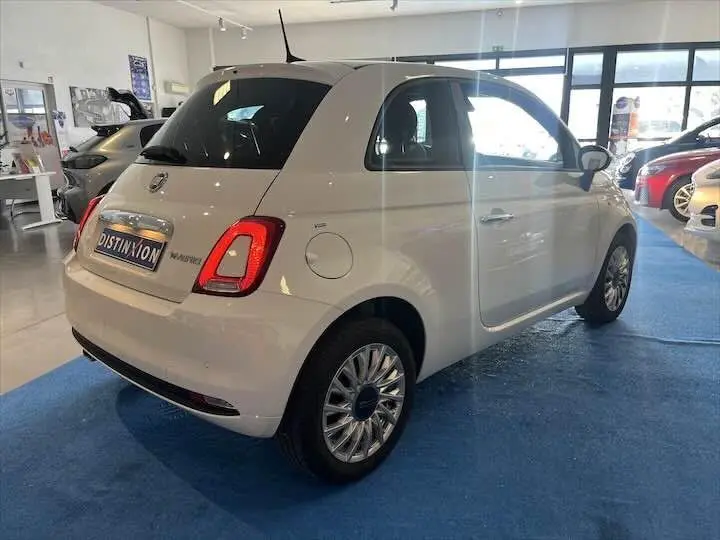 Vue 3/4 arrière droite d'une FIAT 500 blanche 2023 en showroom, avec jantes multibranches et vitres teintées.