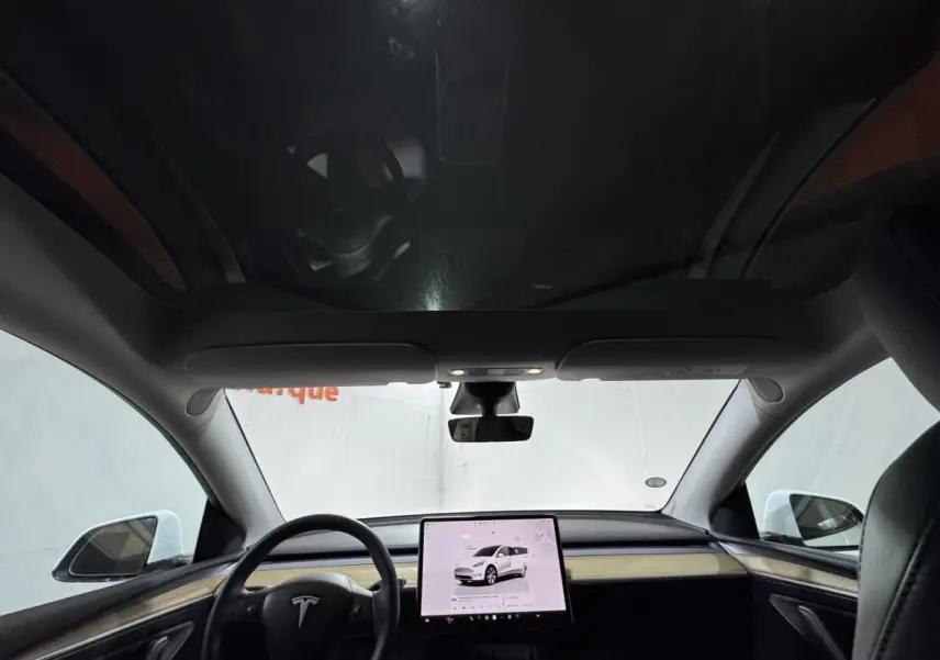 Vue intérieure frontale du tableau de bord et écran tactile central d'une Tesla Model Y blanche avec toit panoramique.