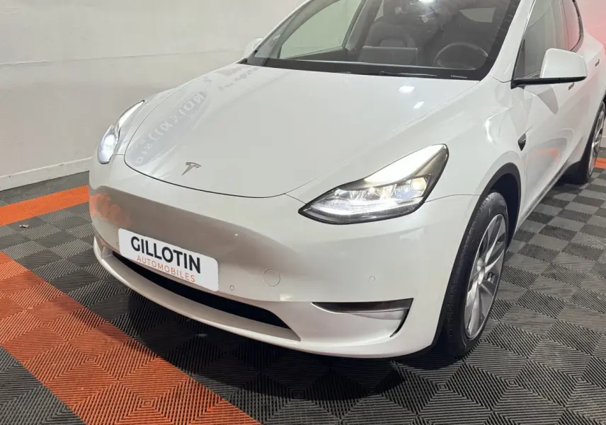 Vue 3/4 avant droit d'une Tesla Model Y blanche avec phares LED allumés en intérieur showroom.