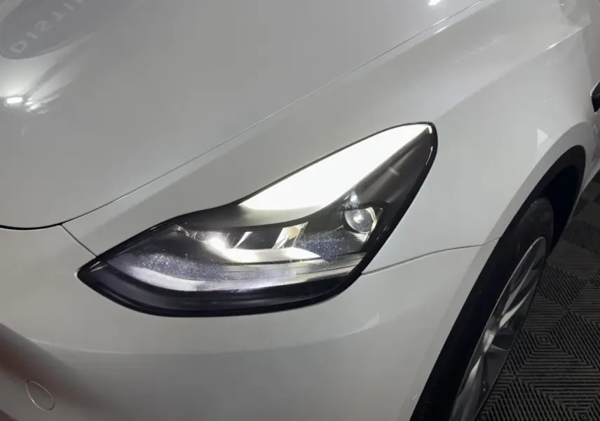 Gros plan sur le phare avant droit allumé d'une Tesla Model Y blanche, mettant en valeur la carrosserie lisse.