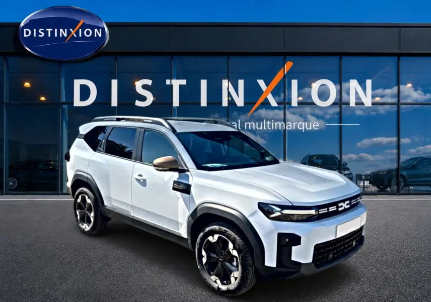 SUV Dacia Bigster blanc en 3/4 avant droit avec jantes noires et barres de toit, devant une vitrine Distinxion.
