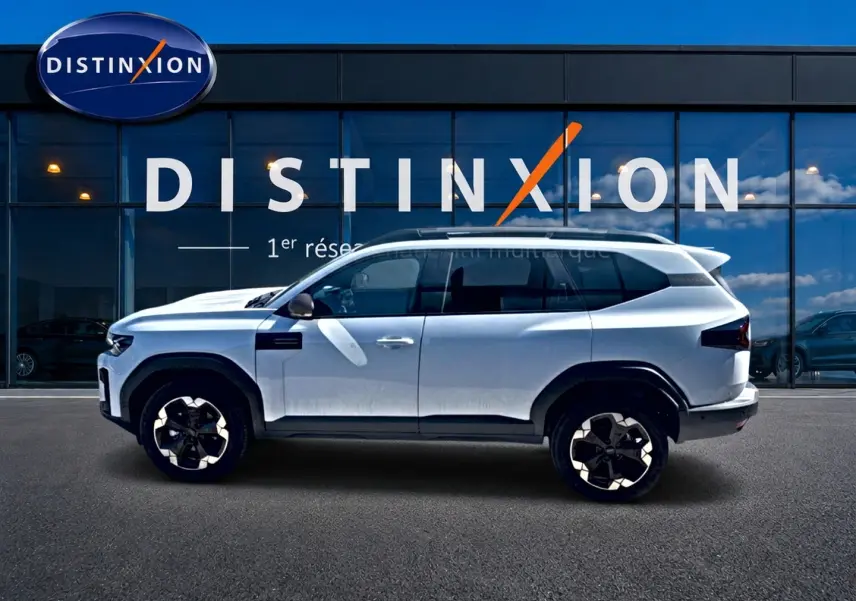 Profil latéral gauche du Dacia Bigster blanc 2026 avec jantes alu et barres de toit devant une vitrine Distinxion.