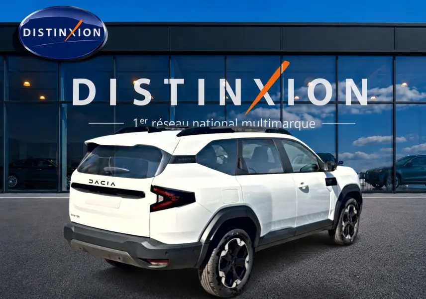 Vue 3/4 arrière droite du Dacia Bigster blanc avec barres de toit et jantes alu noires devant une vitrine Distinxion.