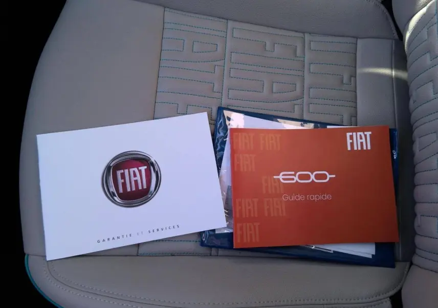 Vue rapprochée des manuels FIAT posés sur le siège avant beige avec surpiqûres bleues du Fiat 600 hybride blanc 2025.