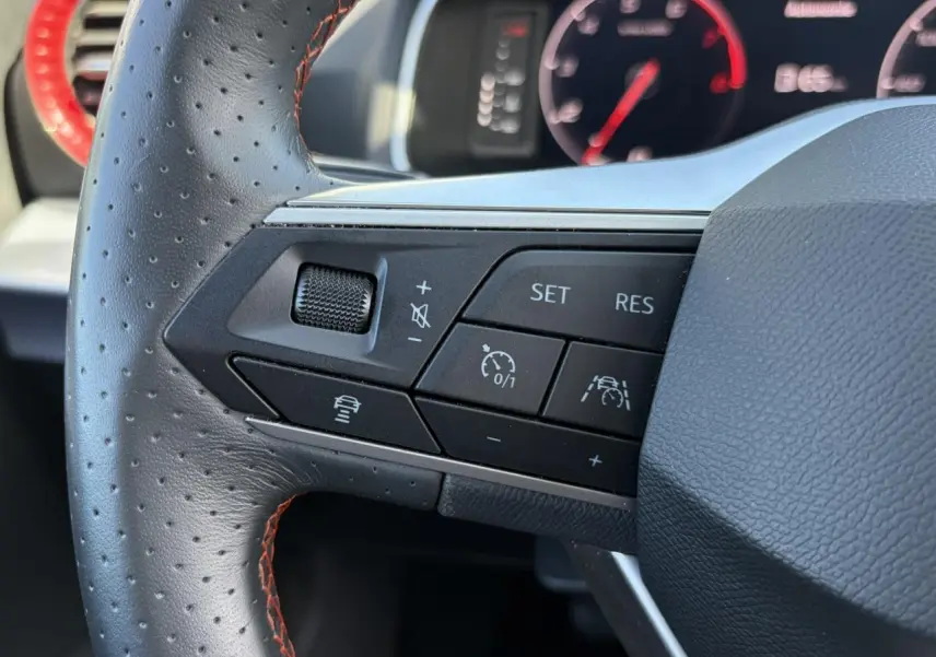 Gros plan sur les commandes à gauche du volant en cuir perforé noir avec surpiqûres rouges du SEAT Ibiza blanc Candy 2025.