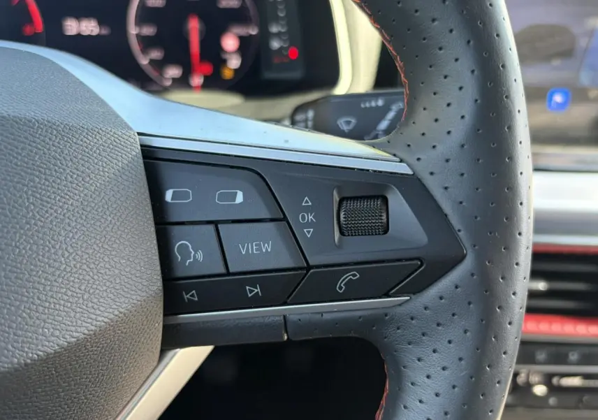 Gros plan sur les commandes gauche du volant perforé d'une SEAT Ibiza blanche, avec tableau de bord numérique en arrière-plan flou.