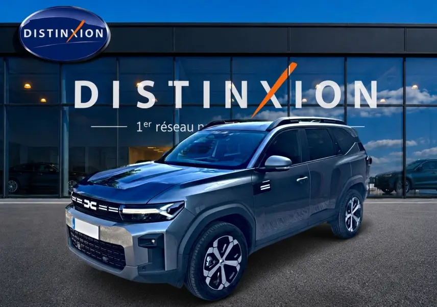 Dacia Bigster gris schiste en 3/4 avant droit, avec jantes alu et barres de toit devant un showroom Distinxion.