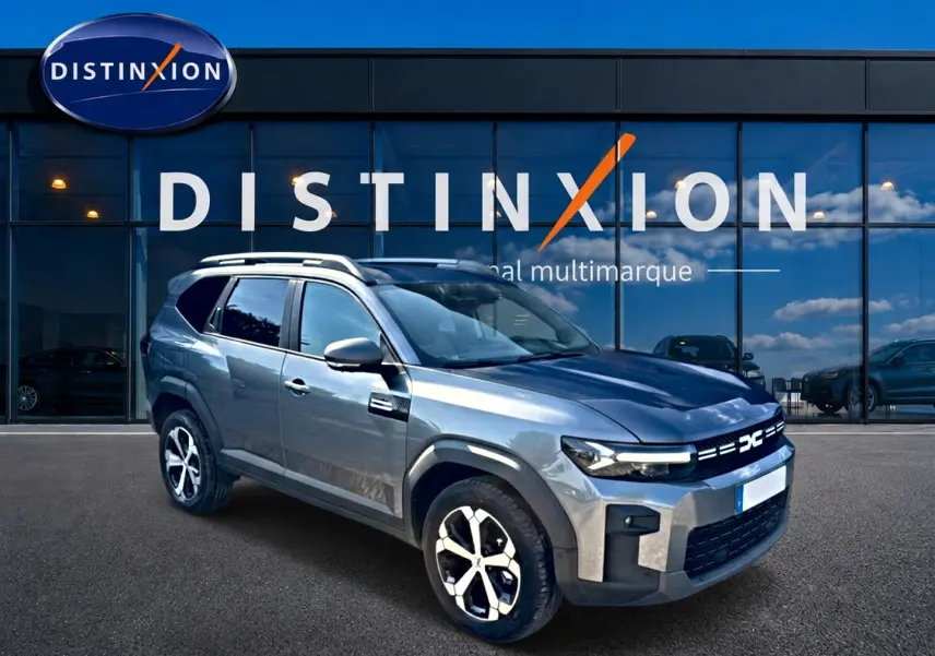 Dacia Bigster gris schiste en 3/4 avant droit, avec barres de toit et jantes alu distinctives, devant un showroom.