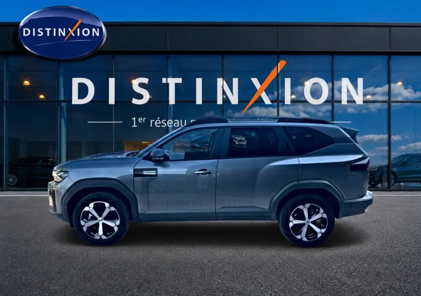 Profil côté gauche du Dacia Bigster gris schiste 2026 avec jantes alu et barres de toit visibles devant une vitrine Distinxion.