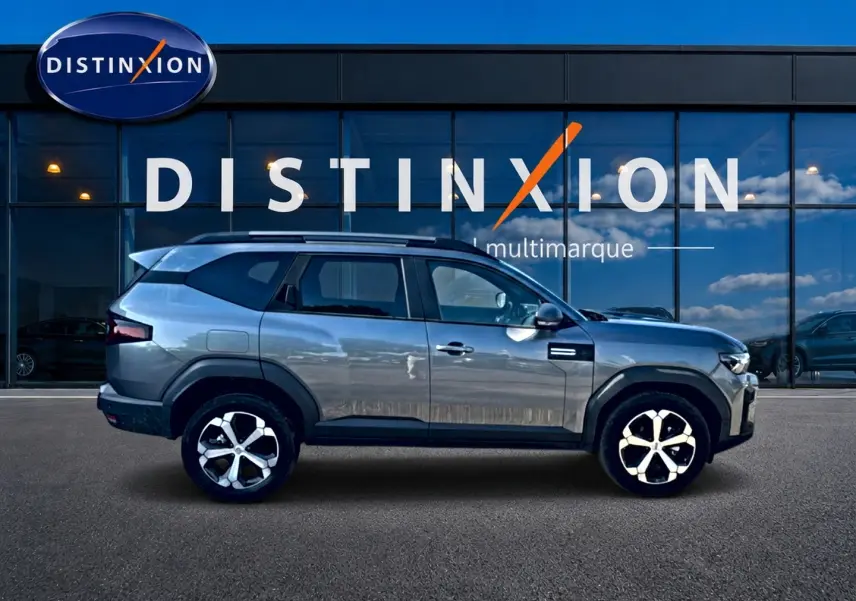 Profil droit du Dacia Bigster gris schiste 2026 avec jantes alu et barres de toit, devant une vitrine Distinxion.