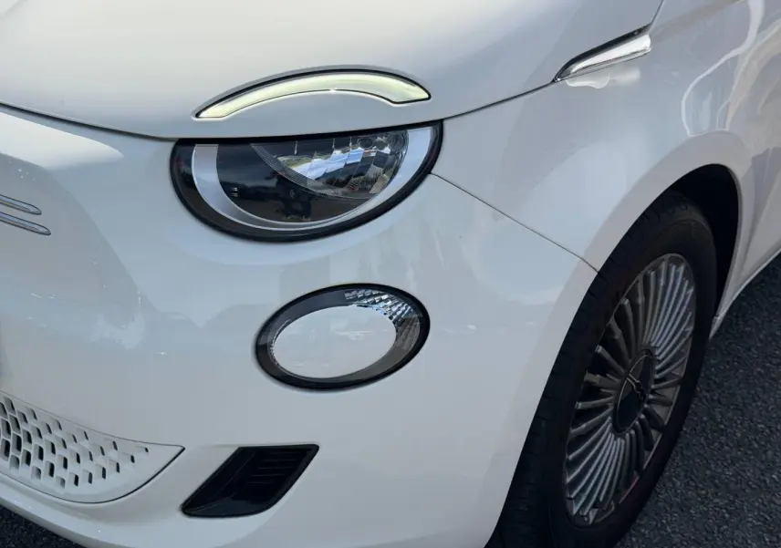 Gros plan sur l'avant droit blanc nacré du Fiat 500 e 118ch MY23, mettant en valeur le phare LED et la jante spécifique.
