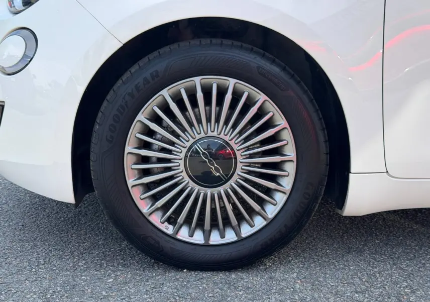 Gros plan sur la roue avant droite d'une Fiat 500 e blanche, avec jante argentée au design rayonné distinctif.