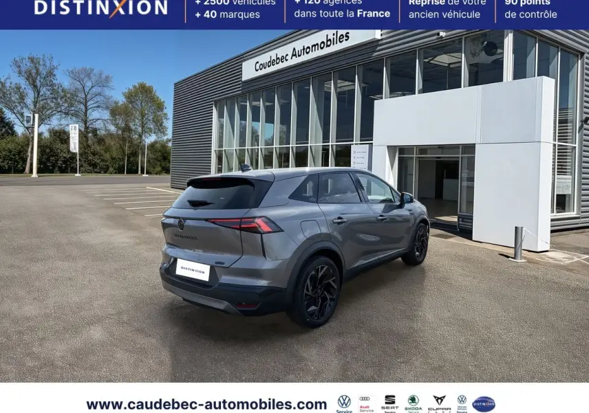 Vue 3/4 arrière droite d’un Renault Symbioz gris métallisé avec jantes noires devant un bâtiment de concession.