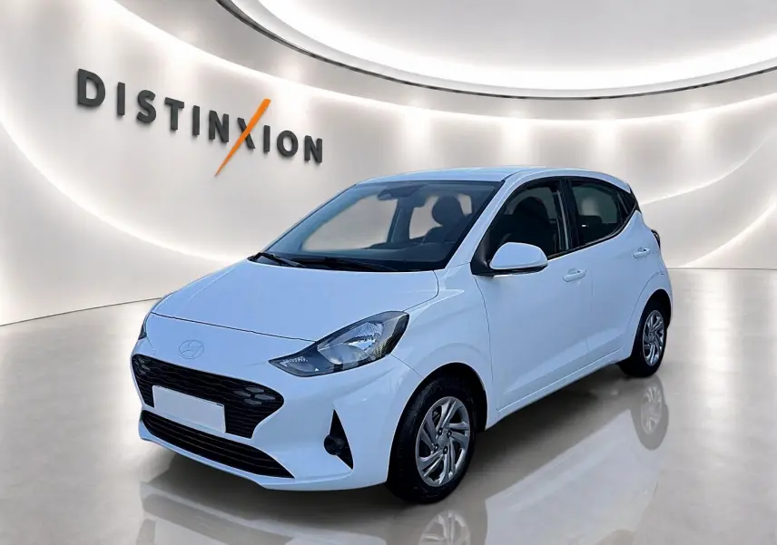 Vue 3/4 avant d'une Hyundai i10 blanche 2024 avec enjoliveurs et rétroviseurs assortis en intérieur showroom lumineux.