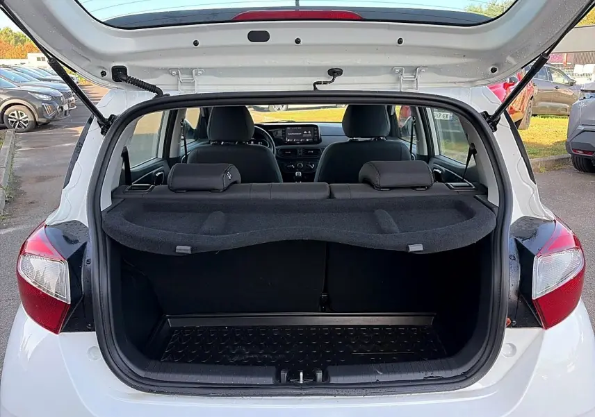 Vue arrière ouverte d'une Hyundai i10 blanche 2024, coffre vide avec tablette cache-bagages et intérieur noir visible.
