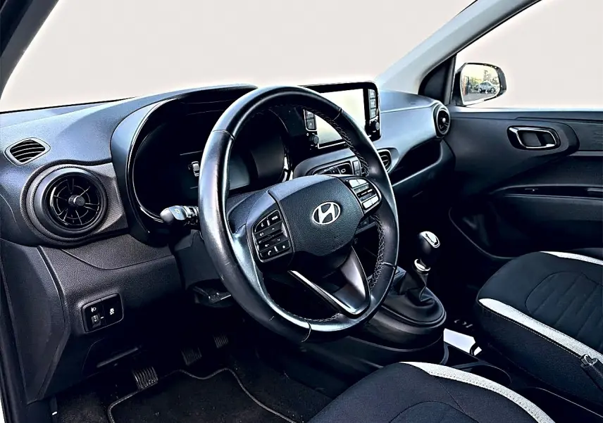 Vue intérieure côté conducteur du tableau de bord et volant cuir noir de la Hyundai i10 2024, avec levier de vitesses manuel.