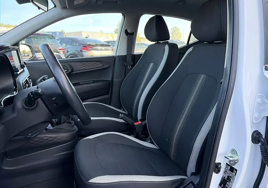 Vue intérieure côté gauche de la Hyundai i10 blanche 2024, montrant les sièges noirs avec surpiqûres grises et volant cuir multifonction.