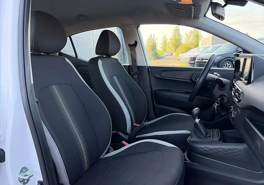 Vue intérieure côté gauche de la Hyundai i10 2024 blanche, montrant les sièges noirs avec surpiqûres grises et la console centrale.
