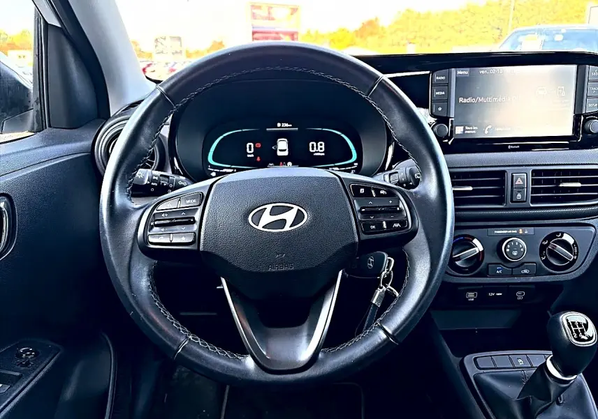 Vue intérieure centrée sur le volant cuir multifonction et le tableau de bord numérique d'une Hyundai i10 blanche 2024.