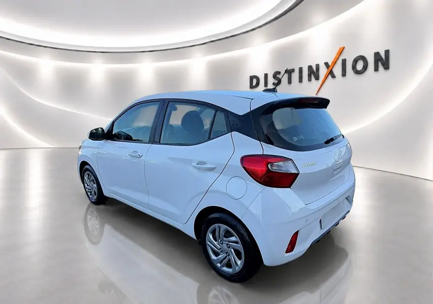 Vue 3/4 arrière droite d'une Hyundai i10 blanche dans un showroom lumineux avec logo Distinxion en arrière-plan.