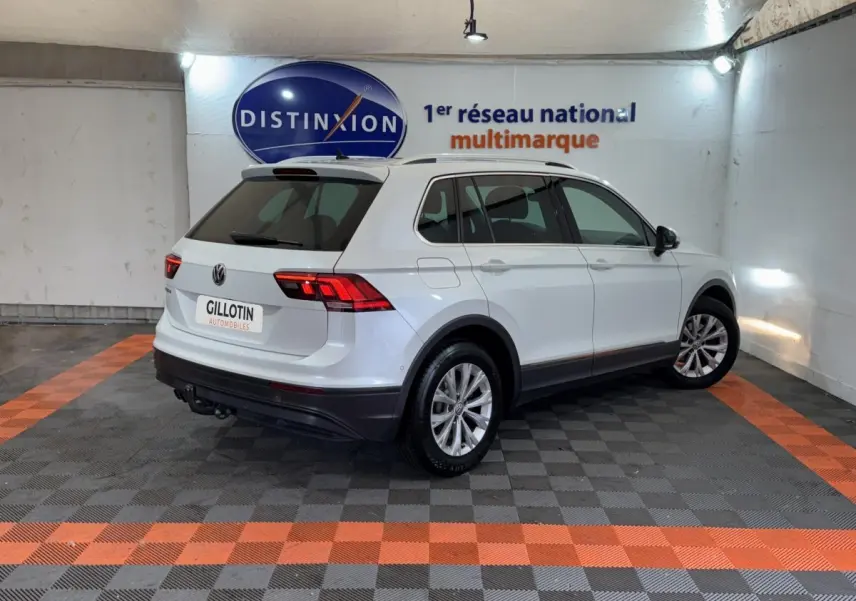Vue 3/4 arrière droite d'un Volkswagen Tiguan blanc stationné en intérieur avec barres de toit noires et jantes argentées.