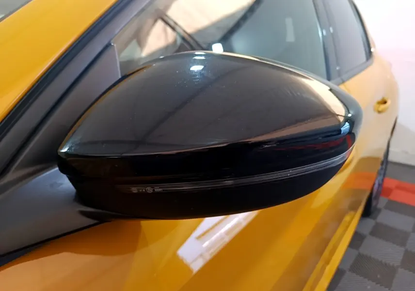 Rétroviseur noir côté gauche en gros plan sur Peugeot 208 jaune vue partielle avant.