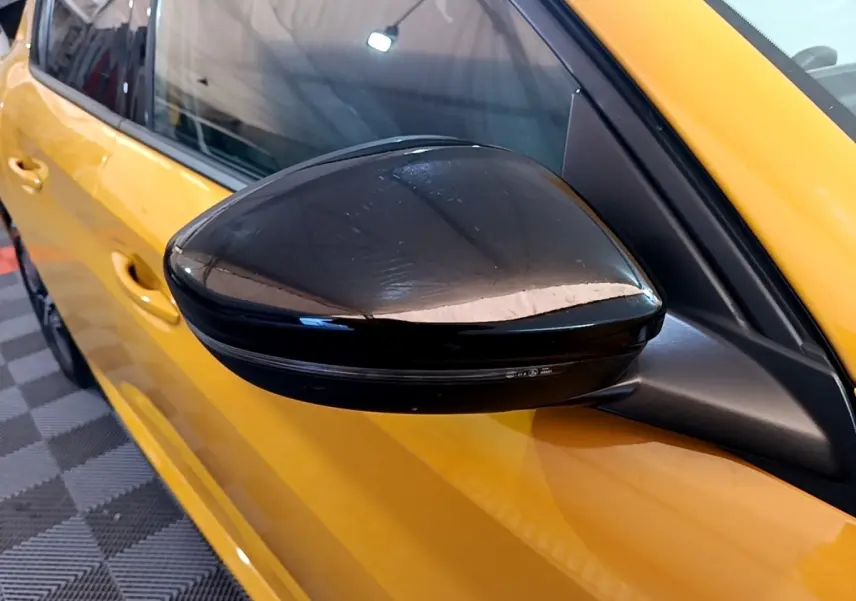 Gros plan sur le rétroviseur noir côté gauche d'une Peugeot 208 jaune vif, avec portière visible en arrière-plan.