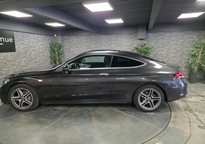 Vue de profil côté gauche d'un coupé Mercedes Classe C gris métallisé avec jantes AMG 18 pouces en intérieur showroom.