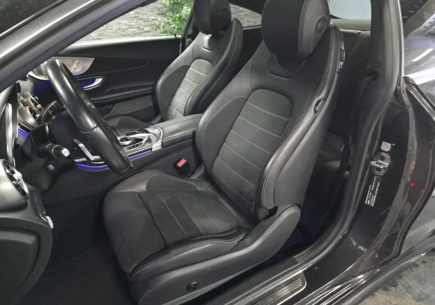 Vue intérieure côté conducteur de la Mercedes Classe C Coupé gris, sièges sport noirs avec surpiqûres blanches et éclairage d'ambiance bleu.