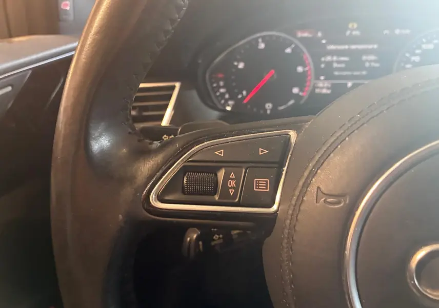 Gros plan sur le côté gauche du volant en cuir noir de l’Audi A8 2016 avec commandes intégrées et tableau de bord flou en arrière-plan.