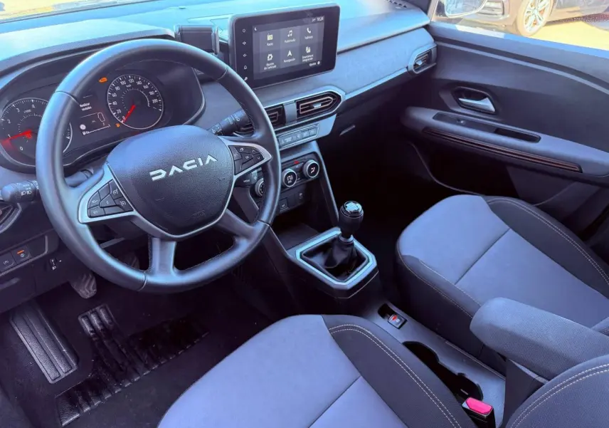 Intérieur de la Dacia Sandero 2025 vu côté conducteur, volant multifonction, écran tactile et sièges tissu brun cuivré.