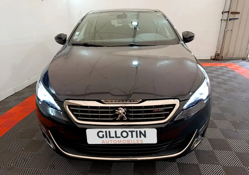 Vue avant d’une Peugeot 308 gris foncé avec phares allumés et calandre chromée dans un garage.