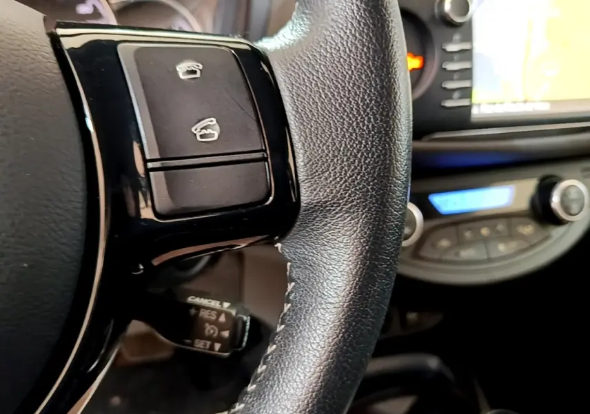 Gros plan sur le volant cuir et alu de la Toyota Yaris blanche avec commandes multifonctions et écran tactile en arrière-plan.