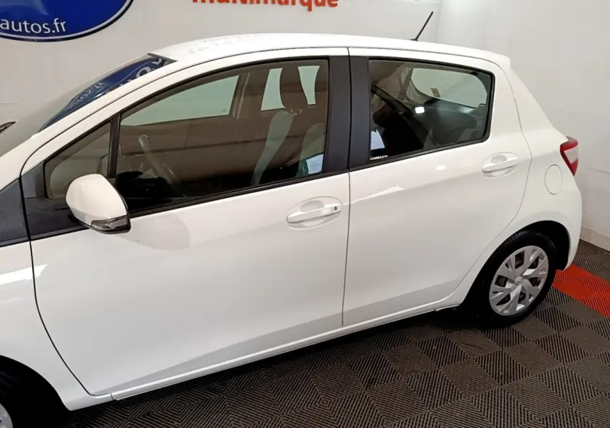 Profil droit d'une Toyota Yaris blanche 2019 en version hybride essence, visible en intérieur sur sol damier noir et rouge.