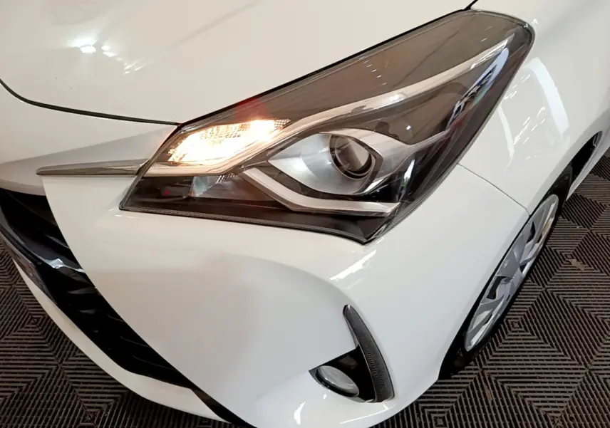 Gros plan sur le phare avant droit allumé d'une Toyota Yaris blanche, avec partie de la calandre et roue visible.