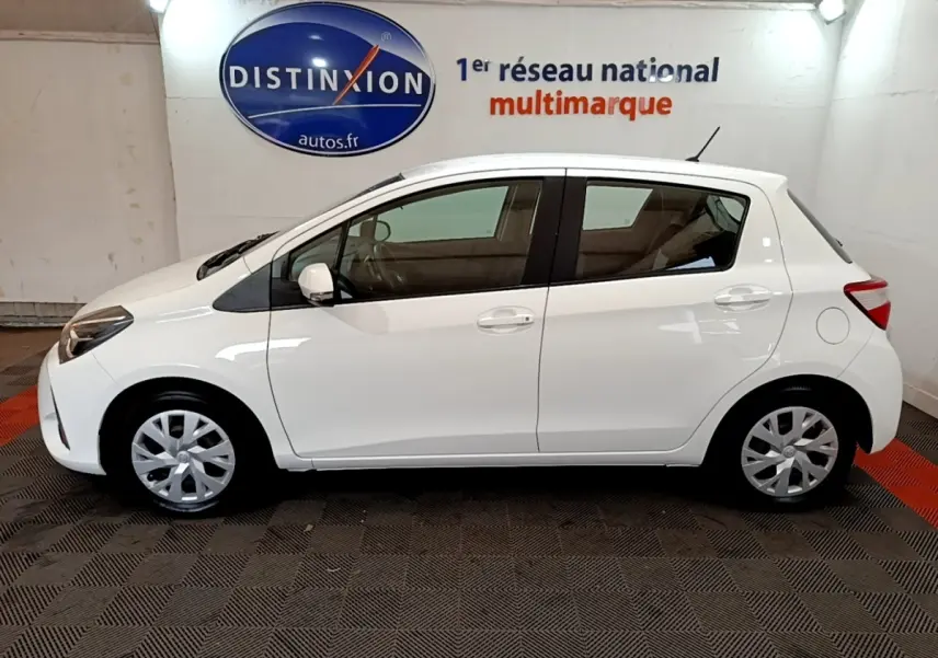 Profil côté gauche d'une Toyota Yaris blanche 2019, compacte, avec jantes alliage et arrière arrondi, en intérieur showroom.