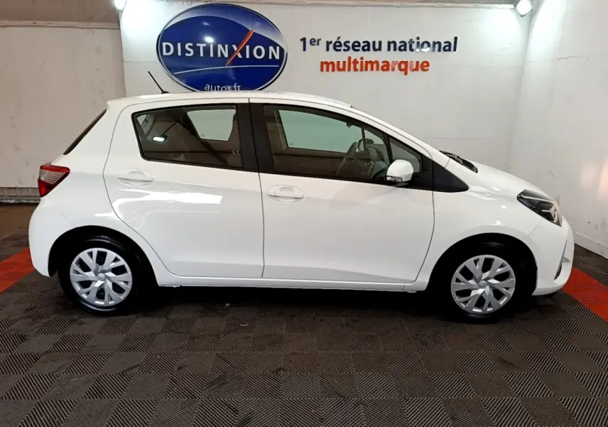 Profil droit d'une Toyota Yaris blanche 2019 essence hybride, vue en intérieur showroom avec sol à motifs.