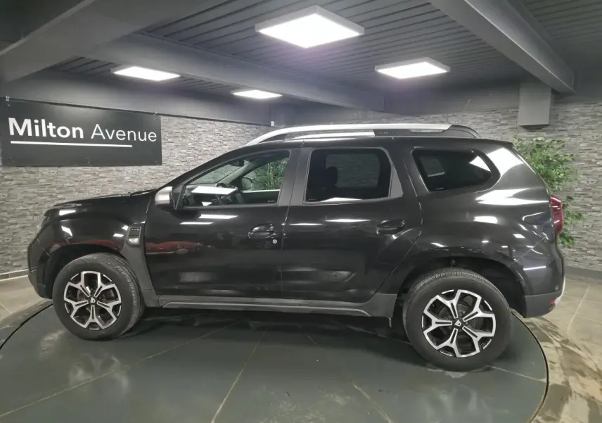 Profil droit du Dacia Duster noir nacré 2019 avec jantes alliage 17 pouces et barres de toit visibles en intérieur showroom.