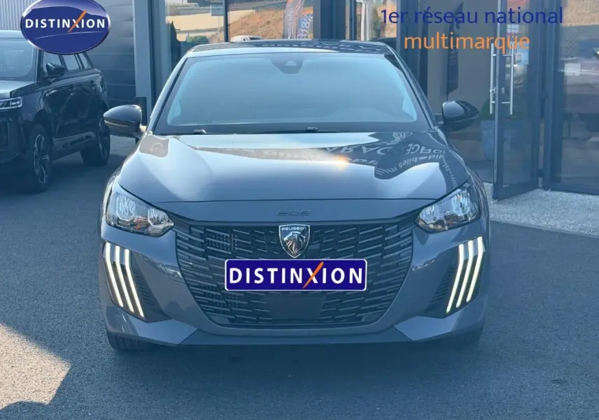 Vue frontale d'une Peugeot 208 1.2 100ch gris Selenium avec feux LED verticaux allumés, stationnée devant un bâtiment.