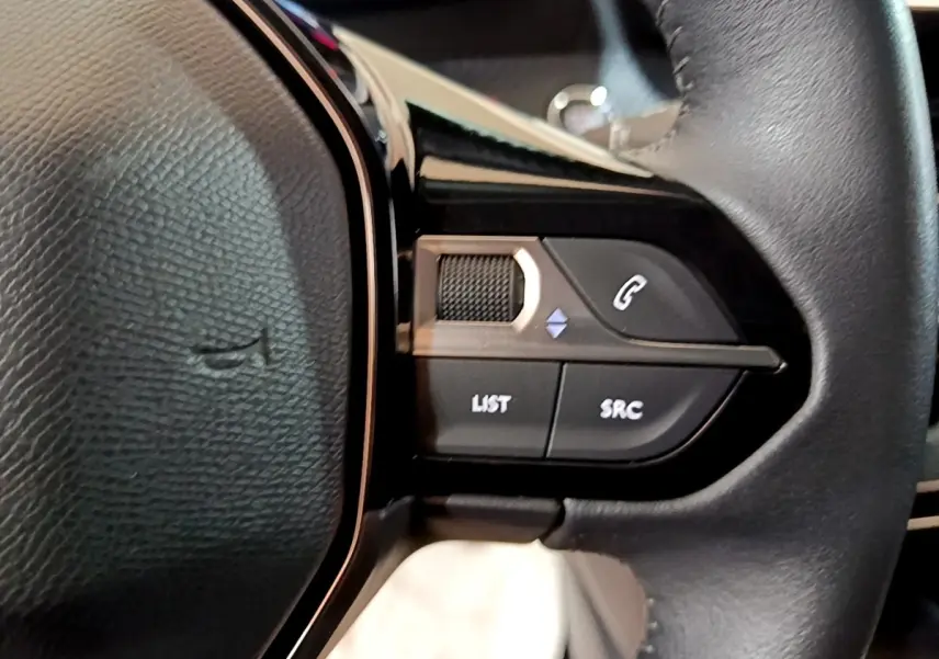 Gros plan sur les commandes multifonctions côté gauche du volant en cuir noir de la Peugeot 208 blanche.