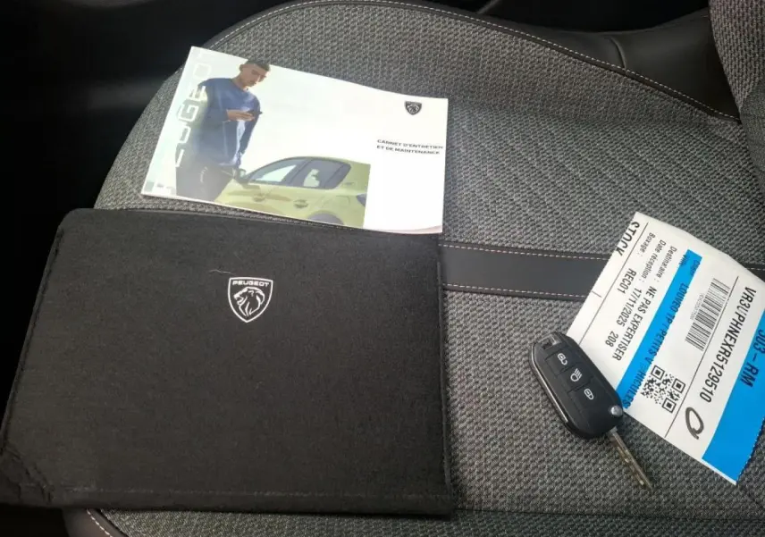 Clé de voiture Peugeot 208 posée sur un siège gris clair avec manuel d'entretien et pochette noire Peugeot.