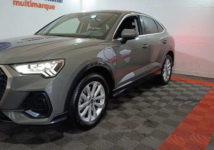Audi Q3 Sportback gris foncé en 3/4 avant droit, phare allumé et jantes alliage visibles dans un showroom.
