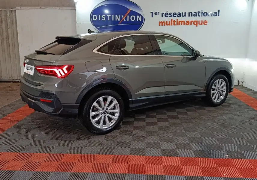 Audi Q3 Sportback gris foncé vue 3/4 arrière droit, avec feux arrière allumés dans un showroom.