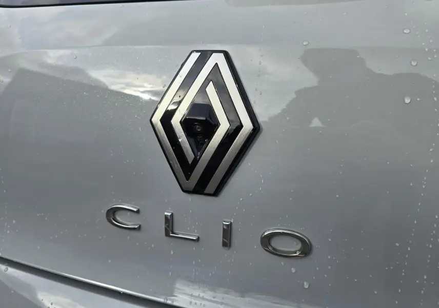 Gros plan sur le logo Renault et le badge Clio sur la malle arrière gris rafale métal avec gouttes d'eau visibles.